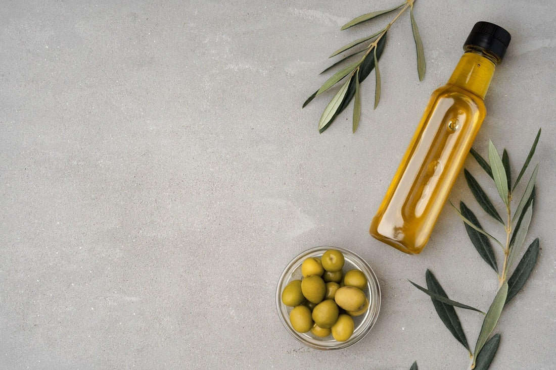 Glass bottle of olive oil with green olives and olive branches on gray background